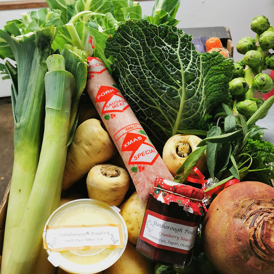 A styled flat-lay of fresh groceries and food items arranged together, including a large leek, a head of cabbage, Brussels sprouts, a jar of Strawberry Sauce, a tub of Buttercream Custard, and a roll of Christmas Special ham from stanborough farm